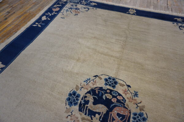 Light beige rug with a dark blue border and a medallion patterned with deer and flowers on wood flooring.