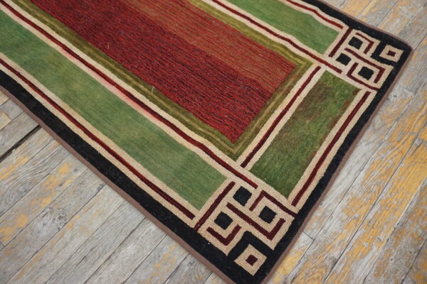 Textured red and green rug with geometric borders rests upon rustic, peeling light wood floorboards.