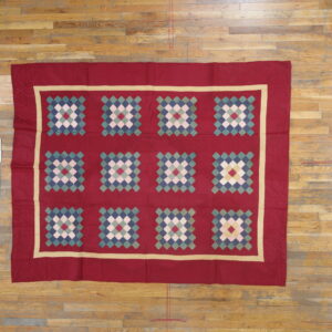 Flat quilted red rug with a tan frame border and colorful diamond grid motifs on natural wood flooring.