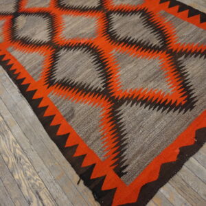 Flatweave rug with a serrated geometric pattern in gray, bright orange, and dark brown on rustic wood flooring.
