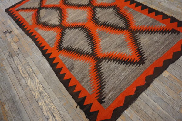 Flatweave rug with a serrated geometric pattern in gray, bright orange, and dark brown on rustic wood flooring.