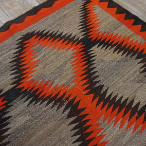 Flatweave rug featuring serrated orange and black diamonds on a natural gray background, resting on weathered wood floors.