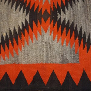 Flat woven rug texture featuring serrated triangles and zigzag patterns in orange, black, and gray.