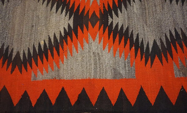 Flat woven rug texture featuring serrated triangles and zigzag patterns in orange, black, and gray.