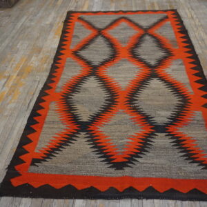 Gray flatweave rug featuring a bold orange and black geometric zigzag diamond pattern on wood flooring.
