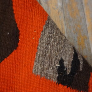 A rug corner featuring bold orange, dark brown, and speckled gray geometric blocks on worn wood flooring.