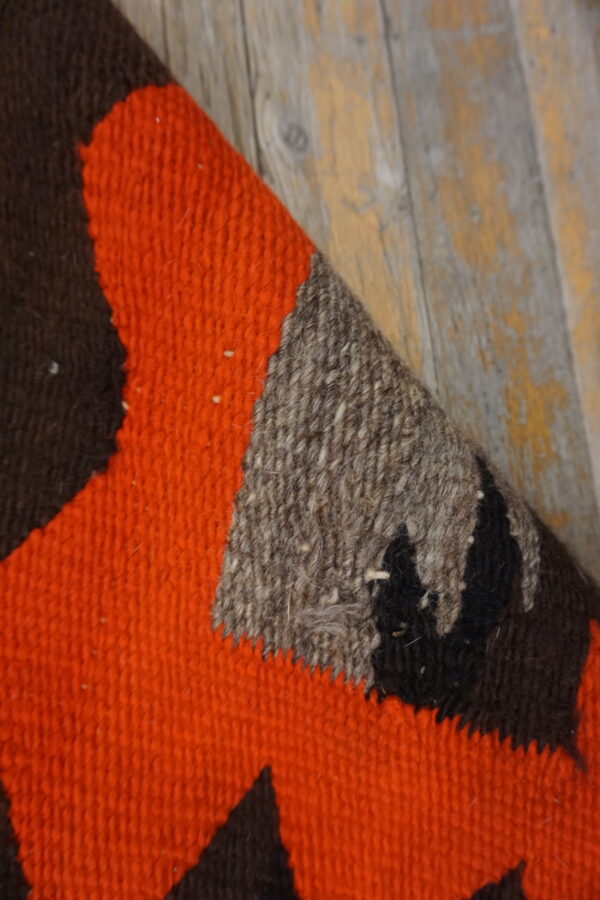 A rug corner featuring bold orange, dark brown, and speckled gray geometric blocks on worn wood flooring.