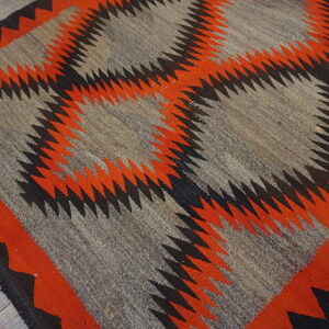 Gray, orange, and black geometric flatweave rug with serrated diamonds resting on light wooden plank flooring.