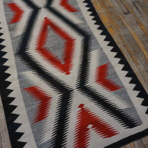 Flat woven rug in red, black, gray, and cream geometric diamond patterns over worn wood flooring.