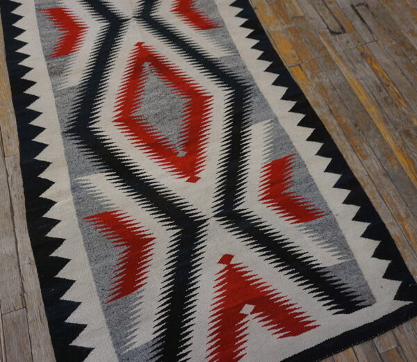 Flat woven rug in red, black, gray, and cream geometric diamond patterns over worn wood flooring.