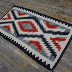 Flatweave rug featuring bold serrated diamonds in red, black, gray, and cream on distressed wood flooring.