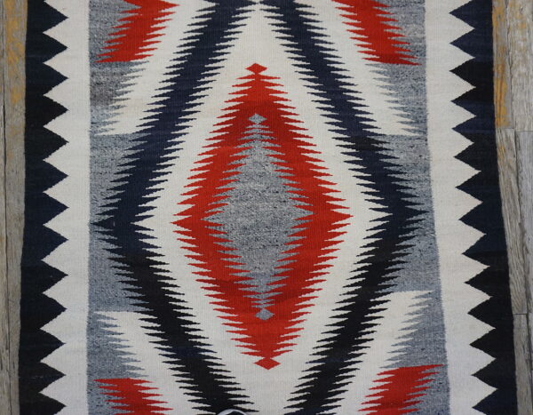 Low-pile rug with interlocking red, black, grey, and white serrated diamond pattern on wood planks.