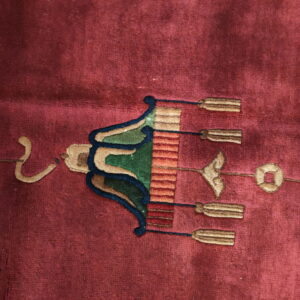 Close-up of a deep red rug showing a stylized green, blue, and tan lantern motif with tassels.
