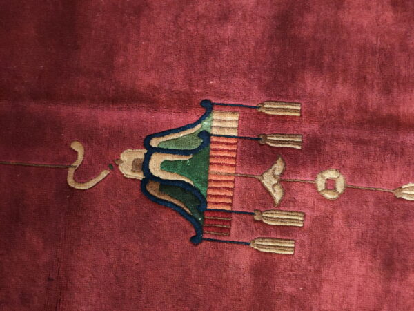 Close-up of a deep red rug showing a stylized green, blue, and tan lantern motif with tassels.