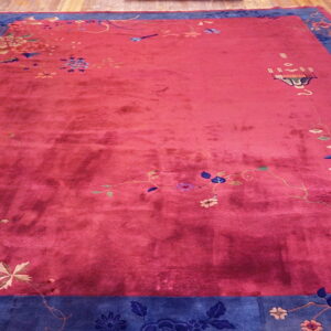A deep pink rug with a wide blue floral border sits on light hardwood flooring.