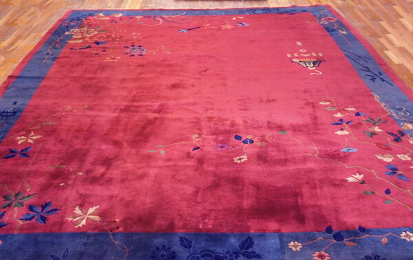 A deep pink rug with a wide blue floral border sits on light hardwood flooring.