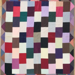 Patchwork textile featuring columns of multicolored rectangular blocks and a triangular border on wood floor.