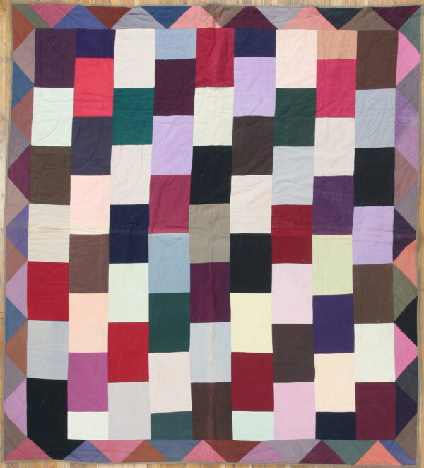Patchwork textile featuring columns of multicolored rectangular blocks and a triangular border on wood floor.