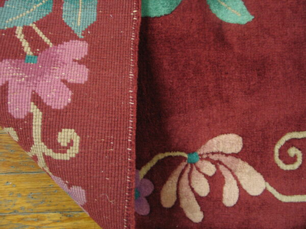 Deep maroon rug with pink and teal flowers is folded, showing the woven reverse side on wood.