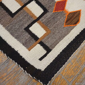 Geometric flatweave rug featuring black, cream, and earth tones, accented by orange details on weathered wood flooring.