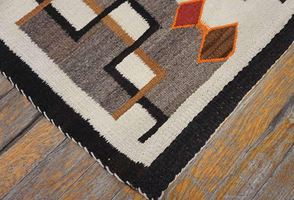 Geometric flatweave rug featuring black, cream, and earth tones, accented by orange details on weathered wood flooring.