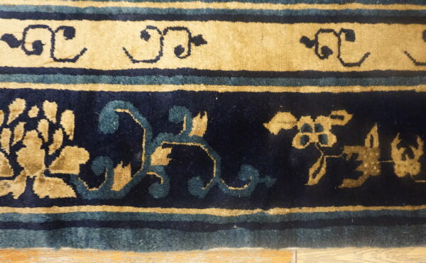Detailed view of a navy and tan rug border with teal stripes and stylized floral motifs on a wood floor.
