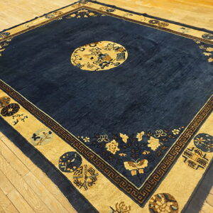 Deep blue square area rug with elaborate tan and gold borders lying on light wooden floorboards.