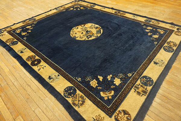 Deep blue square area rug with elaborate tan and gold borders lying on light wooden floorboards.