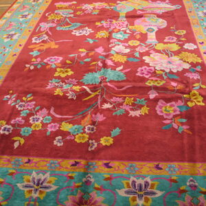 Plush red rug with multicolored floral design, turquoise border, and intricate vase motifs on wood floor.