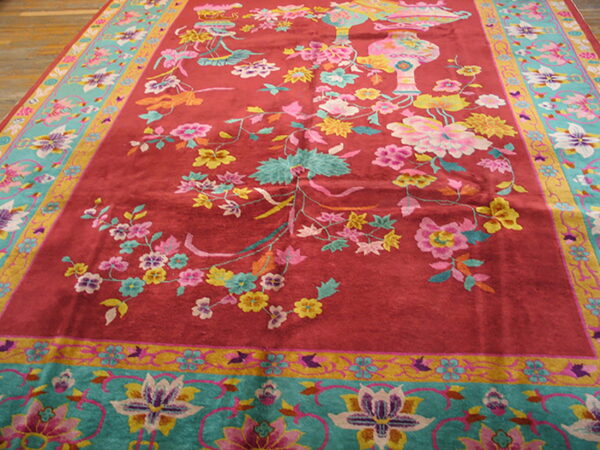 Plush red rug with multicolored floral design, turquoise border, and intricate vase motifs on wood floor.