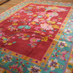 Reddish-pink rug with multicolor floral designs and a wide teal and gold border rests on hardwood planks.