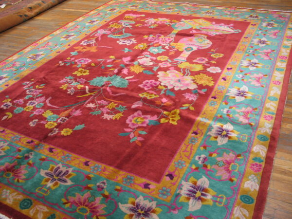 Reddish-pink rug with multicolor floral designs and a wide teal and gold border rests on hardwood planks.