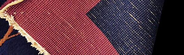 The reverse side of a rug showing woven maroon and speckled navy blue fabric blocks and a beige edge.