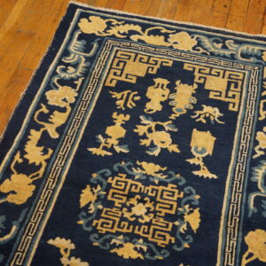 Low-pile navy blue rug featuring yellow and teal traditional patterns, including a geometric key border, rests on wood flooring.