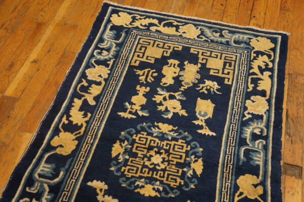 Low-pile navy blue rug featuring yellow and teal traditional patterns, including a geometric key border, rests on wood flooring.