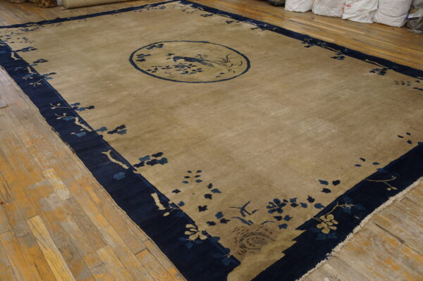A tan rug with a dark blue border and central bird medallion rests on worn hardwood flooring.