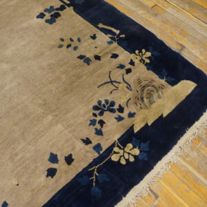 Tan rug with navy blue floral border and visible fringe rests on worn yellow plank flooring.