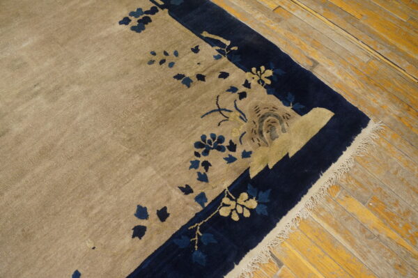 Tan rug with navy blue floral border and visible fringe rests on worn yellow plank flooring.