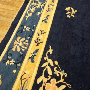 Navy blue rug with gold and teal floral borders rests on light brown hardwood flooring.