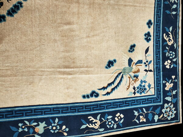 Beige rug corner with a navy blue border featuring fretwork, floral vines, and a stylized blue bird.