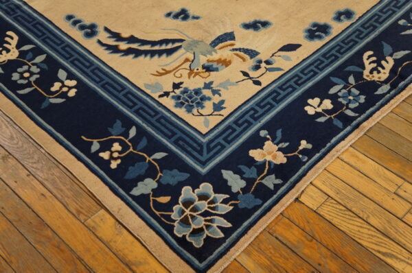 Tan rug corner with blue phoenix and floral designs, detailed navy border, resting on shiny hardwood planks.