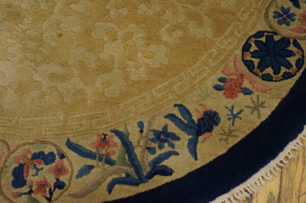 A curved beige rug section showing tone-on-tone texture, navy border, colorful blue and pink floral accents, and fringe.