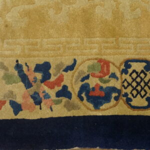 Gold rug close-up showing a geometric key border and a colorful band of floral and knot medallions.