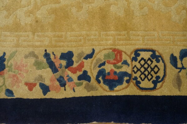 Gold rug close-up showing a geometric key border and a colorful band of floral and knot medallions.