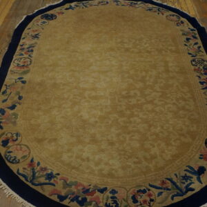 Oval tan rug featuring a sculpted center and a multicolored floral border on distressed wood flooring.