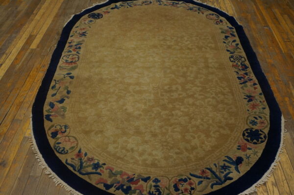 Oval tan rug featuring a sculpted center and a multicolored floral border on distressed wood flooring.