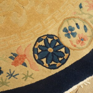 A golden tan rug displays a navy border and stylized coral and blue floral medallions on wood flooring.