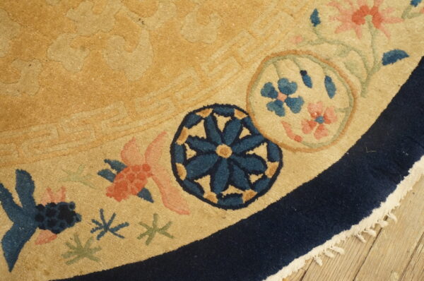 A golden tan rug displays a navy border and stylized coral and blue floral medallions on wood flooring.