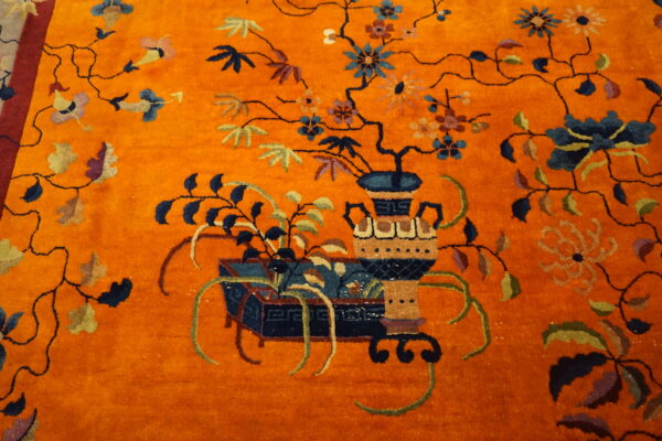 Orange high-pile rug detail showing a decorative vase and vines with blue, green, and multicolored flowers.