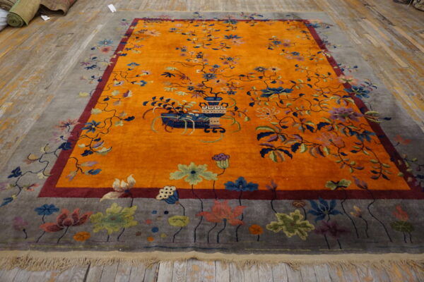 Saffron orange rug featuring a large multicolored floral vase design and a gray pictorial border on wood floor.